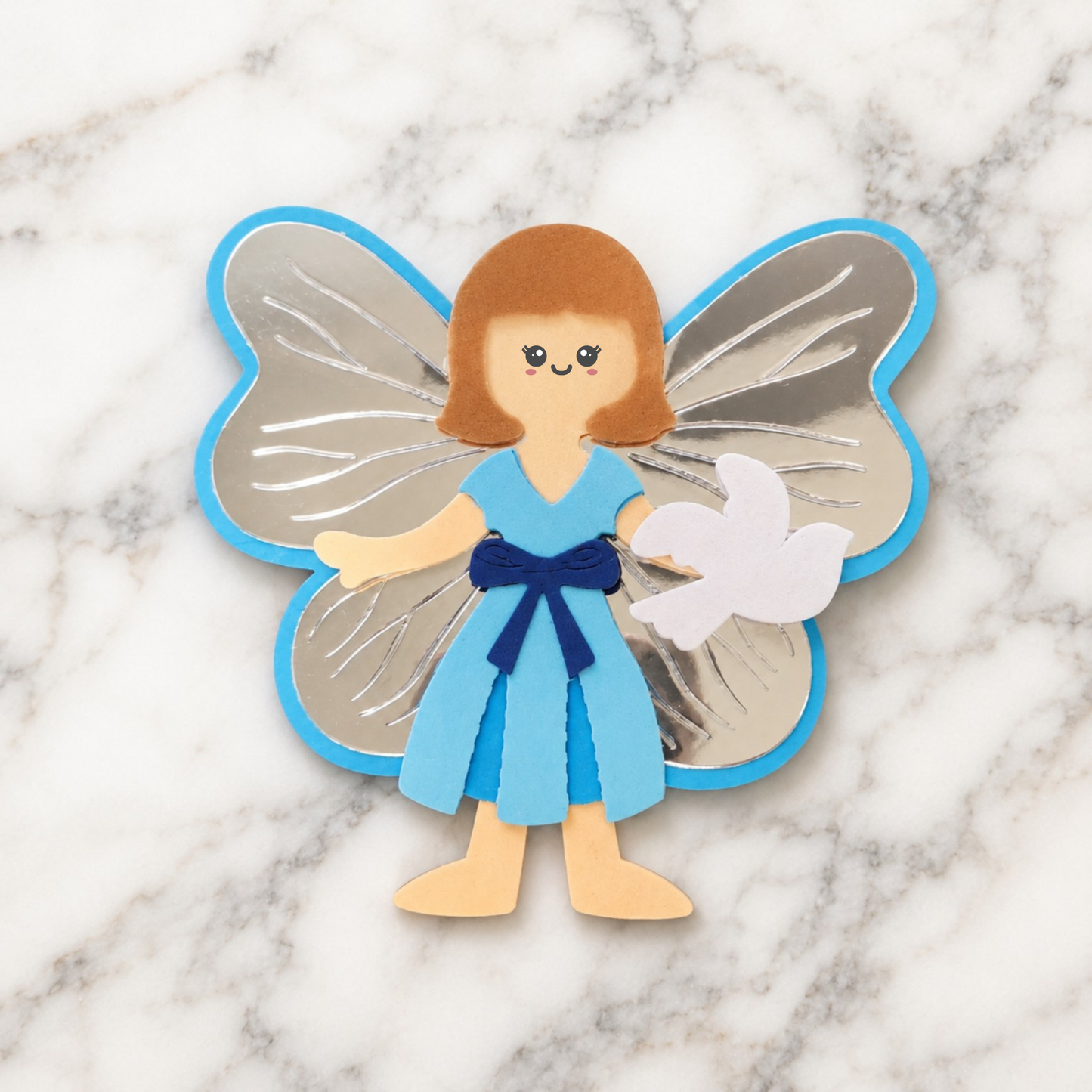 Handmade Christian Peace Fairy paper craft with brown hair, light blue dress, dark blue sash and metallic silver wings displayed on a white and grey marble background.